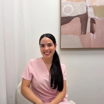 A smiling woman in a pink dress sitting on a chair indoors.
