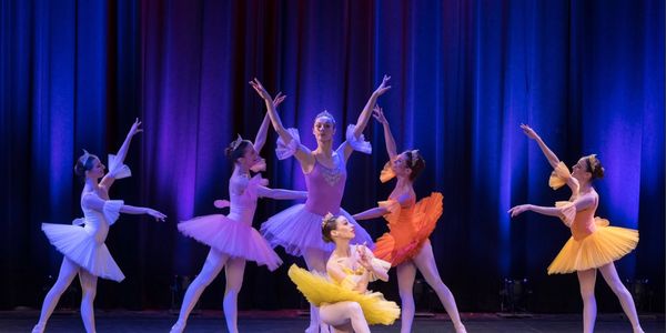 Ballet dancers in colorful tutus perform gracefully on stage with blue curtains.