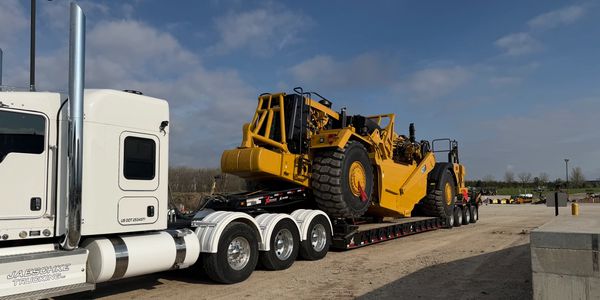 White truck hauling large yellow construction equipment on a flatbed trailer.