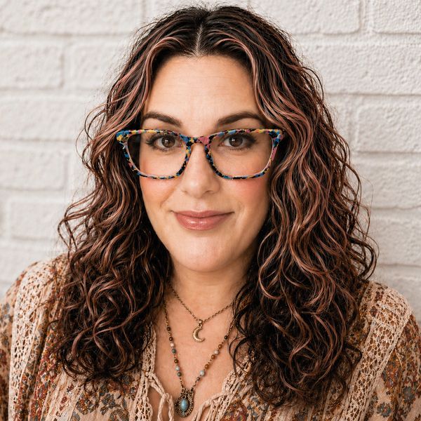 Smiling woman with curly hair and colorful glasses against a white brick wall.