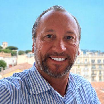 Smiling man in striped shirt outdoors with city backdrop.