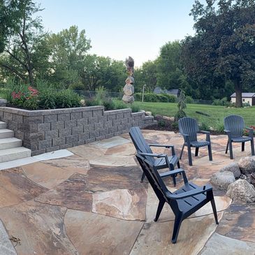 A retaining wall with stairs descending to a slate rock patio and firepit area. 