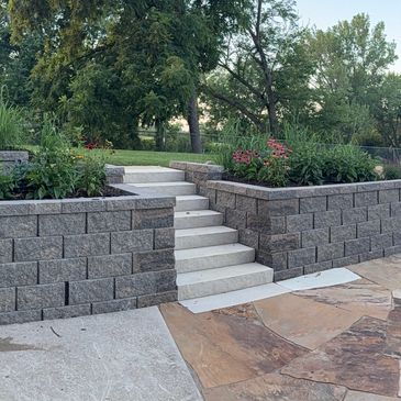 A retaining wall with stairs descending to a slate rock patio and firepit area. 
