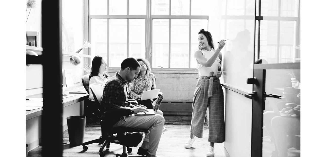 A woman leads a discussion at a whiteboard with colleagues in a bright office.