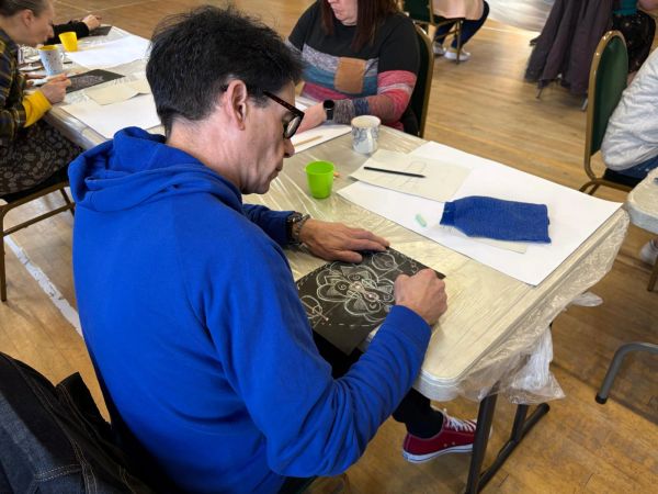 Man in blue hoodie drawing on black paper at a table.