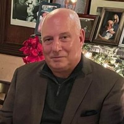 A middle-aged man in a dark jacket smiling indoors with framed photos and red flowers in the background.