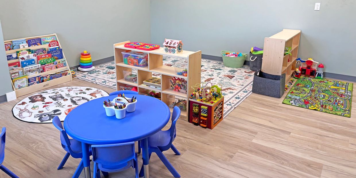 Tidy daycare room with toys, play mats, table, chairs, and bookshelf.