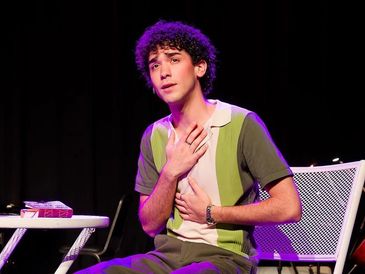 A young man expresses emotion while seated on stage under purple lighting.