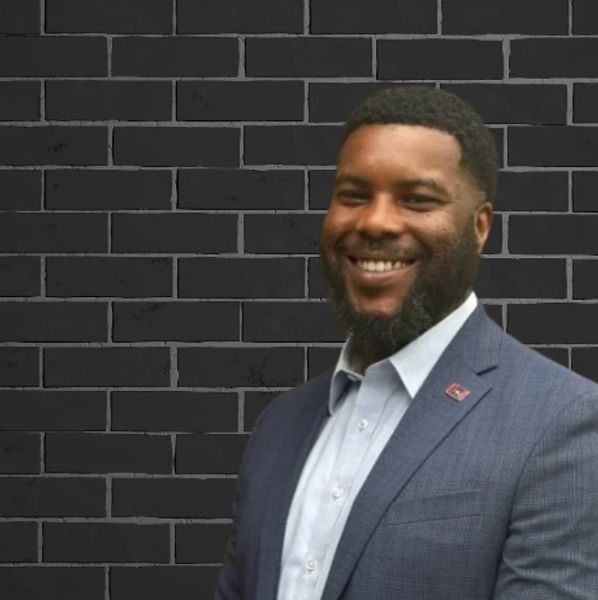 Smiling man in a blue suit stands against a dark brick wall.