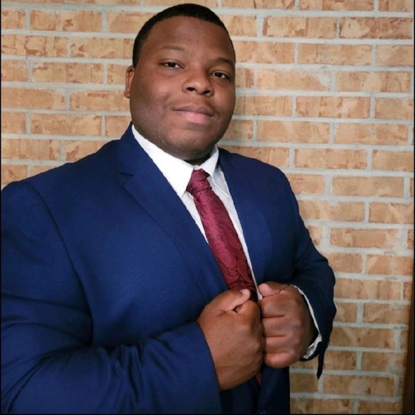 Man in a blue suit and red tie posing confidently against a brick wall.