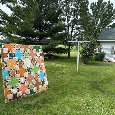 Colorful quilt hanging on a clothesline in a green yard.