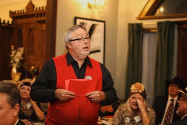 Man in a red vest speaking at a social gathering indoors.