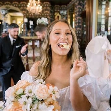 Bride smiling with wedding cake in mouth, holding bouquet.