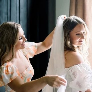 A woman helps another adjust her wedding veil indoors.
