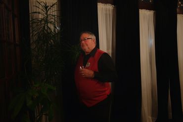 A man in a red vest stands holding a drink indoors near plants and curtains.