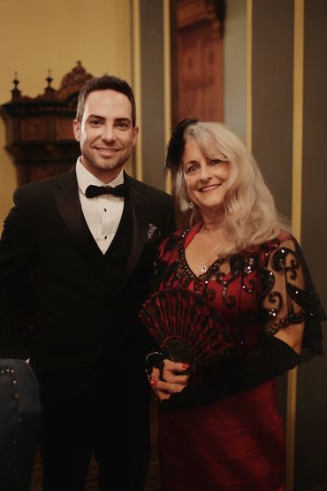 A man in a tuxedo and a woman in a red dress with a fan pose together indoors.