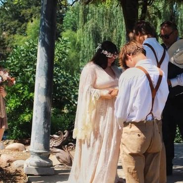 A couple stands in a serene outdoor setting, dressed in vintage-style wedding attire.