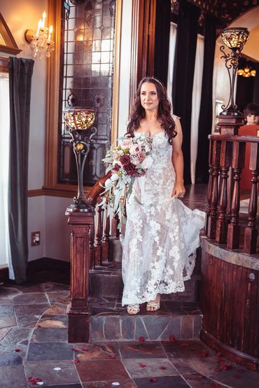 Bride in a lace wedding dress holding a bouquet, descending stairs in an elegant venue.