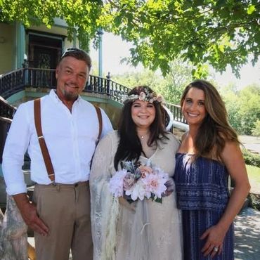 Three people smiling outdoors, one holding flowers in a white dress.