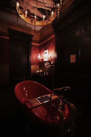 Dimly lit vintage bathroom with a red clawfoot tub and ornate chandelier.