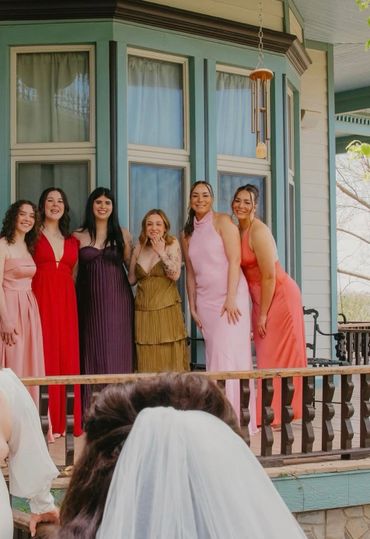 A bride admires six bridesmaids in colorful dresses on a porch.