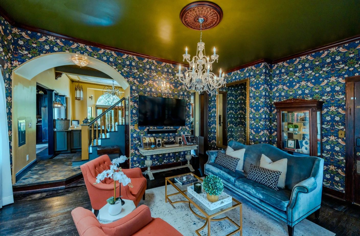 Eclectic living room with floral wallpaper, velvet blue sofa, coral armchairs, and a crystal chandelier.