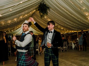 Two men in kilts dancing joyfully under twinkling lights at a festive event.