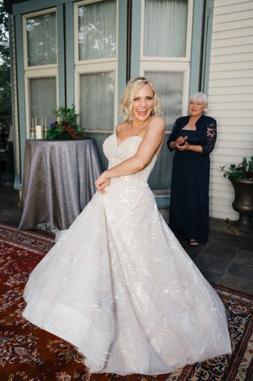 Bride twirling happily in her wedding dress with a smiling guest in the background.