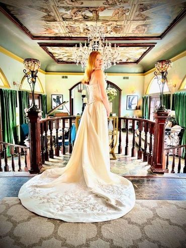 A bride in a flowing gown stands in an elegant, vintage-style room with ornate ceiling art.