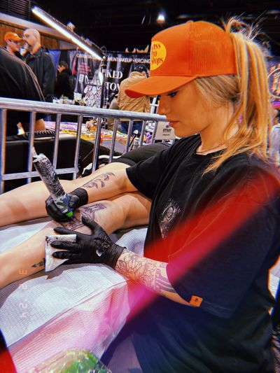 Tattoo artist in an orange cap working on a client's leg tattoo at a convention.