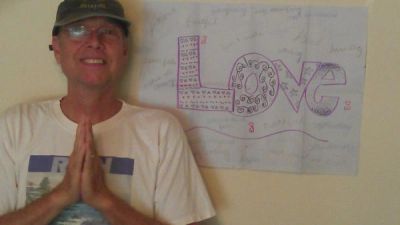 A man with glasses and a cap poses with hands in prayer gesture beside a 'LOVE' sign on the wall.