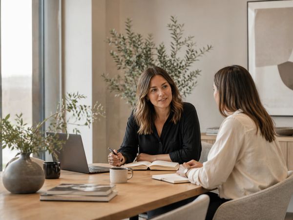 Two women having a discussion at a wooden table with a laptop and notebooks.