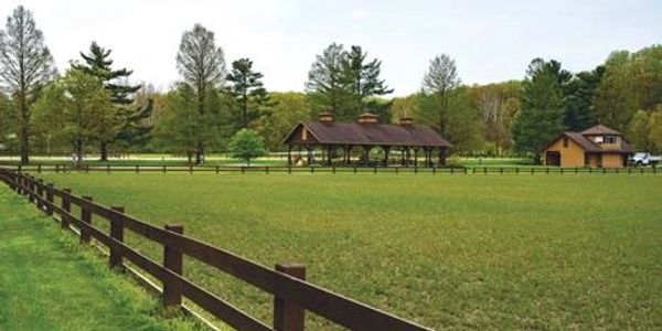Spacious green field enclosed by wooden fences with small buildings and trees in the background.