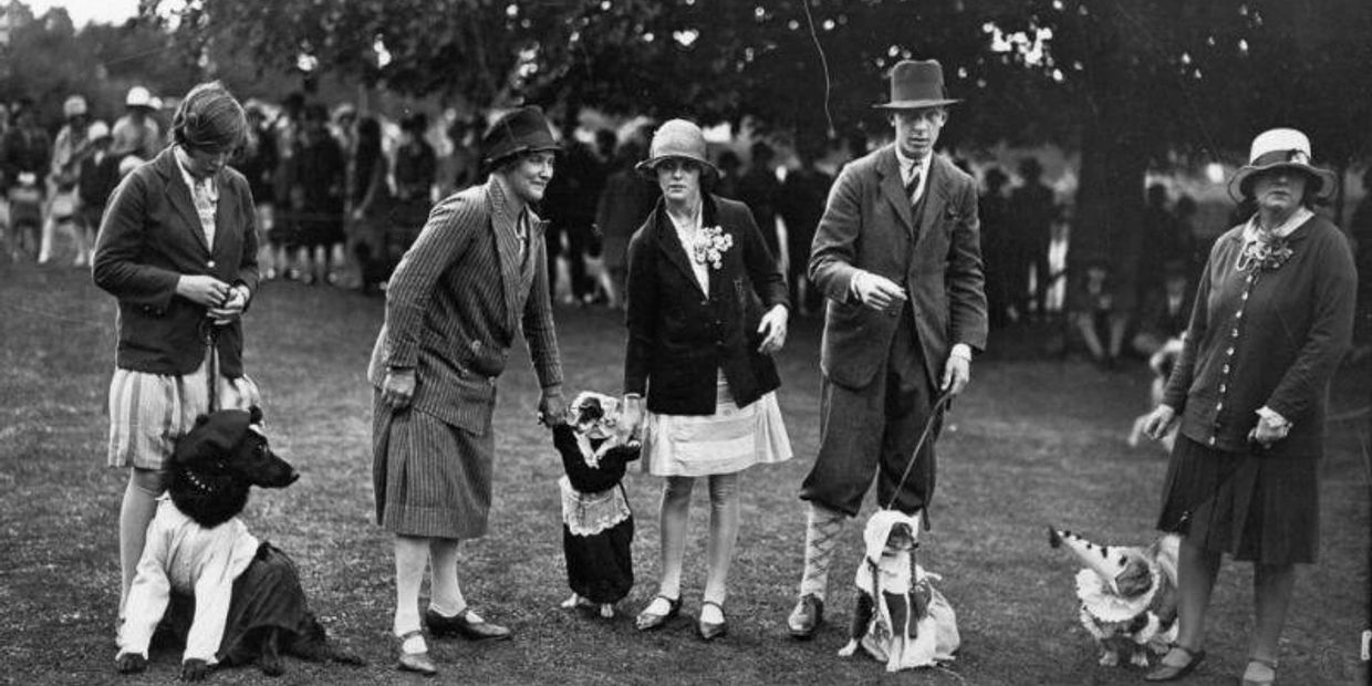 People in 1920s attire with dogs dressed in costumes at an outdoor event.
