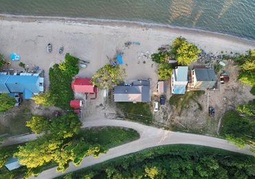 Aerial view of beach houses and boats by the shoreline with trees and a sandy road.