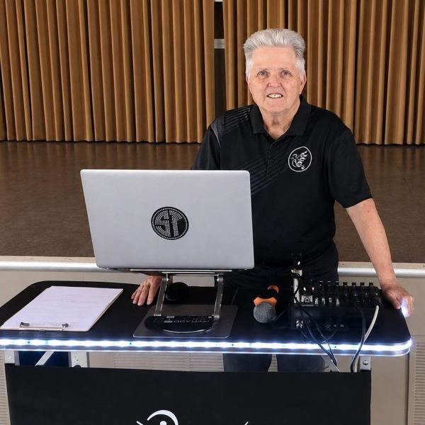 Elderly man standing behind DJ setup with laptop and microphone.
