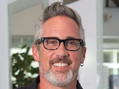 Smiling man with gray hair, glasses, and a beard indoors.