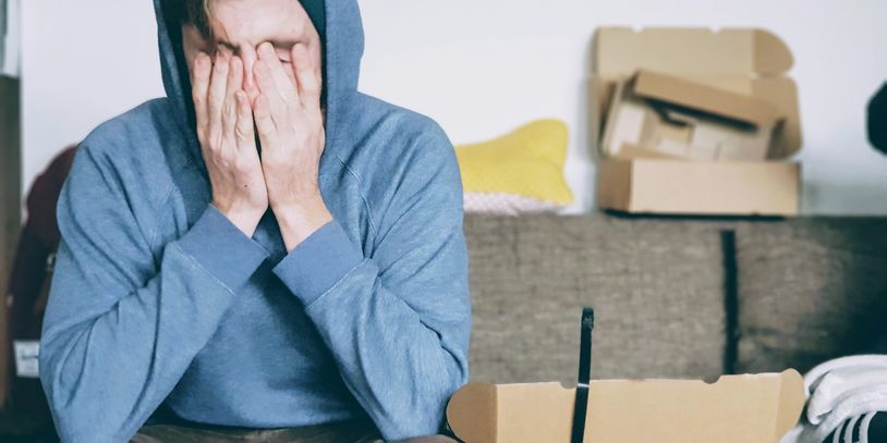 Person in a hoodie covering their face, sitting on a couch with cardboard boxes nearby.
