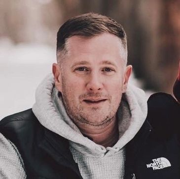 Man in a gray hoodie and black vest outdoors.