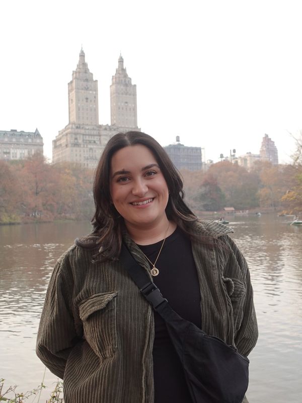 Smiling woman standing by a lake with city buildings in the background.