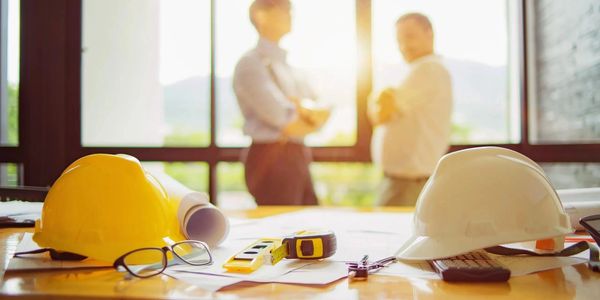 Construction helmets and tools on a table with two professionals talking in the background.
