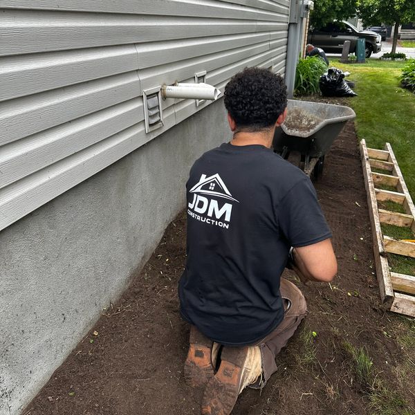 A worker kneeling on soil near a house exterior with a wheelbarrow and ladder.