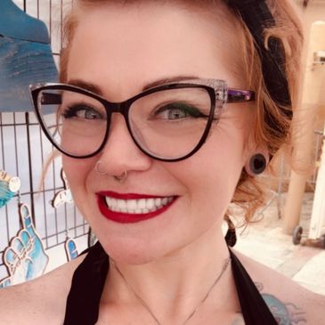 Smiling woman with glasses, red lipstick, and piercings.