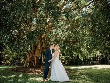 A couple sharing a kiss under a large tree in a sunny park.