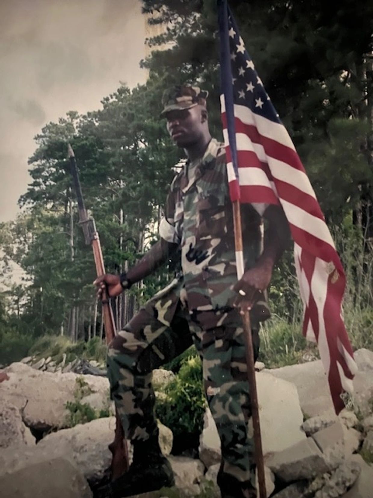 Soldier in camouflage holding an American flag and rifle outdoors.