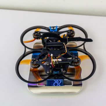 A small drone with propeller guards placed on a digital scale showing 292 grams.