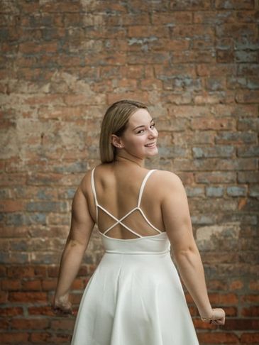 Smiling woman in a white dress poses against a brick wall, showing back details.