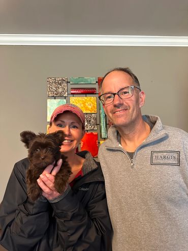 A smiling couple with a small brown dog indoors.