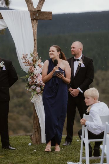Woman in navy dress speaks at outdoor wedding ceremony beside man in tuxedo.
