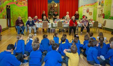 a group of young school children in uniforms sit on the floor opposite a table where a group of olde